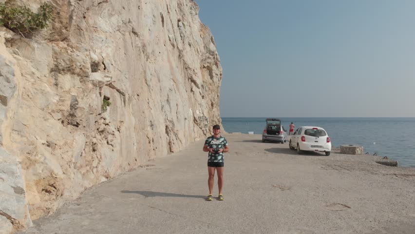 Male tourist controls drone. Aerial panorama drone view of Perissa beach near mountain, black pebble beach. Santorini Island, Greece. Thira city. Aegean Sea, Mediterranean.
