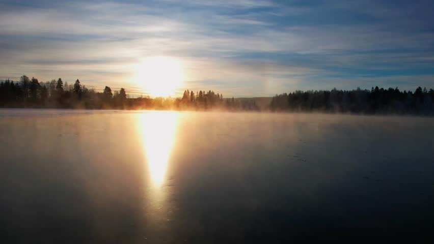 A drone footage of a frozen lake in the woods with a scenic sunset scene in the background
