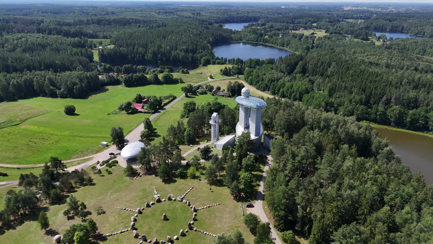 Aerial shot of Moletai Astronomical Observatory in Lithuania, featuring telescopes, a stone circle, and lush green forest under a clear blue sky.