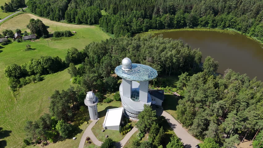 Aerial shot of Moletai Astronomical Observatory in Lithuania, featuring telescopes, a stone circle, and lush green forest under a clear blue sky.