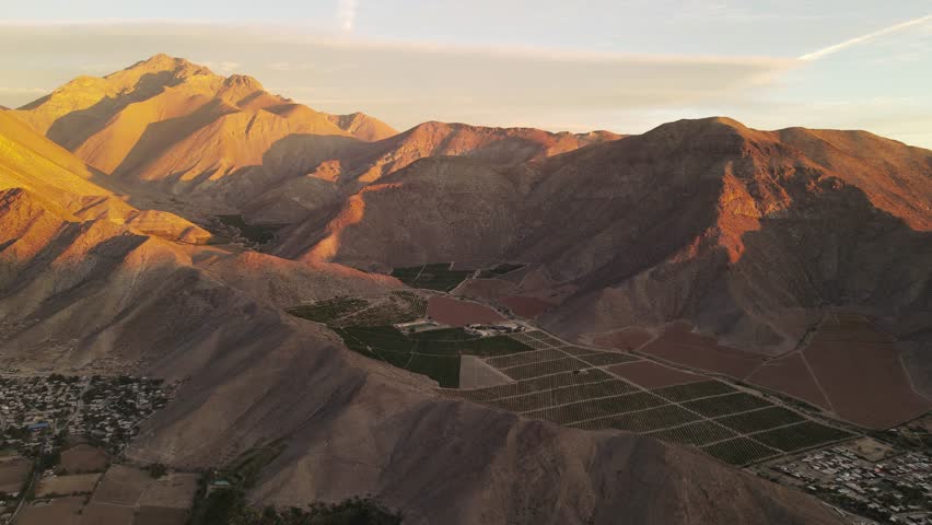A high drone footage of cultivated fields near rural mountainside village with mountains in the background at sunset