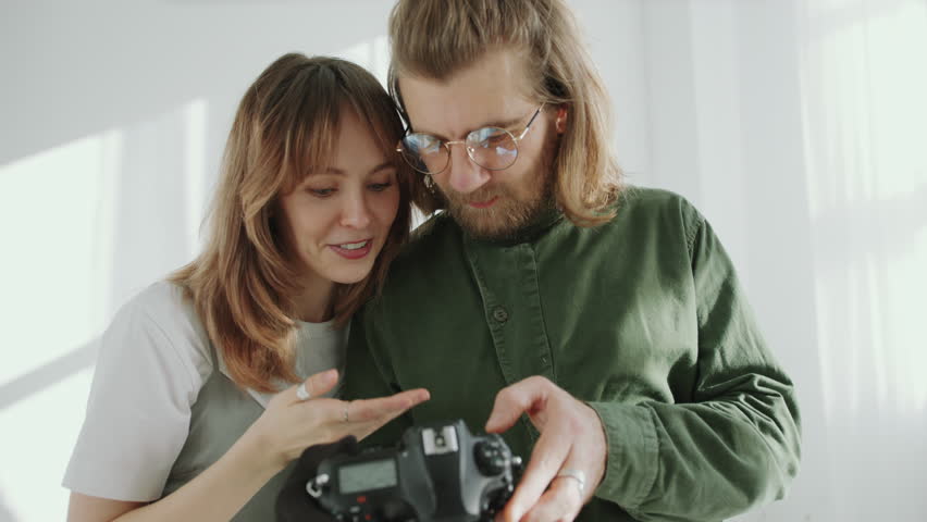Young cheerful woman and photographer examining pictures on camera viewfinder, talking and smiling after photo session in studio