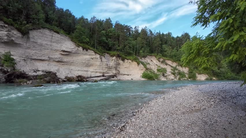 A serene river flowing alongside a rocky shoreline, bordered by lush green trees under a bright blue sky, capturing the peaceful beauty of nature