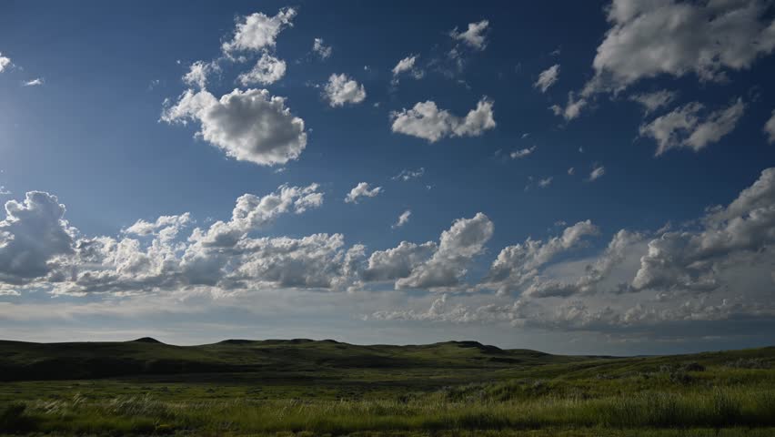 Time lapse of white summer clouds moving above a prairie badlands setting with blowing grass and distant rolling hills.  The sunlight hitting the scene varies as the clouds move.  
