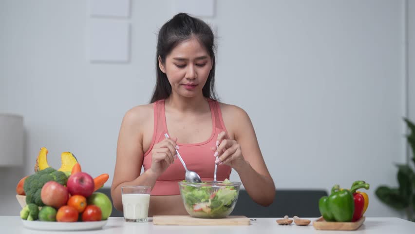 Woman enjoying a fresh salad in a kitchen with vibrant vegetables, promoting a healthy lifestyle