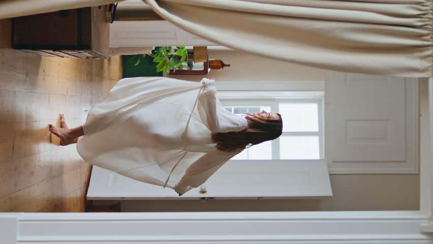 Positive girl relaxing at cozy house alone vertically. Happy woman enjoying weekend morning at light interior. Dancing lady in silk robe celebrating carefree lifestyle at beige apartment. Lazy day