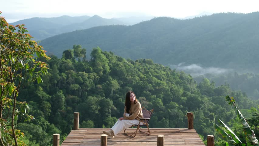 A woman drinking coffee while sitting on a chair and enjoy watching a beautiful mountains view