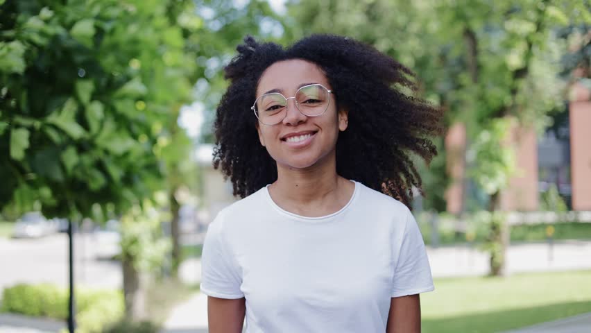 Portrait of charming african american woman wearing clear glasses walking among trees outdoors. Optimistic young woman dressed casually in white t-shirt gazing directly at camera with pleasant smile.