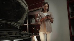 A woman stands in a garage with the hood of a car open, looking down at her phone while holding it in her hands - Powered by Shutterstock - Get 15% off with code: PIKWIZARD15