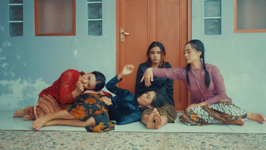 Four women in traditional attire performing a dance in front of a door. One woman is lying on the floor while the others are seated and gesturing with their hands.