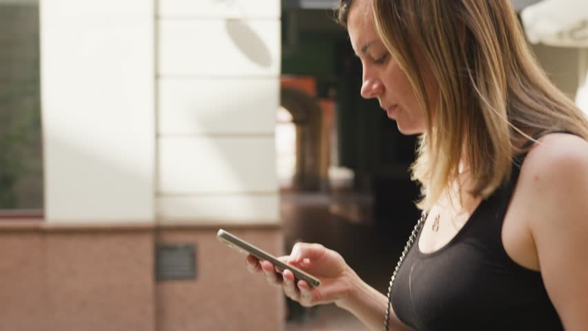 Woman engrossed in her smartphone while walking on bustling city street, surrounded by urban life. Female traveler using mobile phone in European city