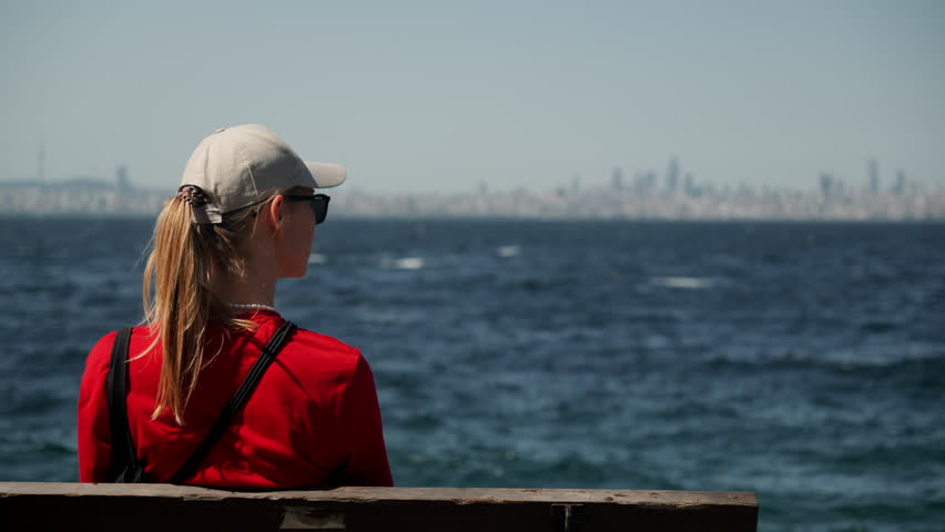 Girl in beige cap and red jacket sits on bench on embankment with bay in sea in background. Young woman in her thoughts on coastline.