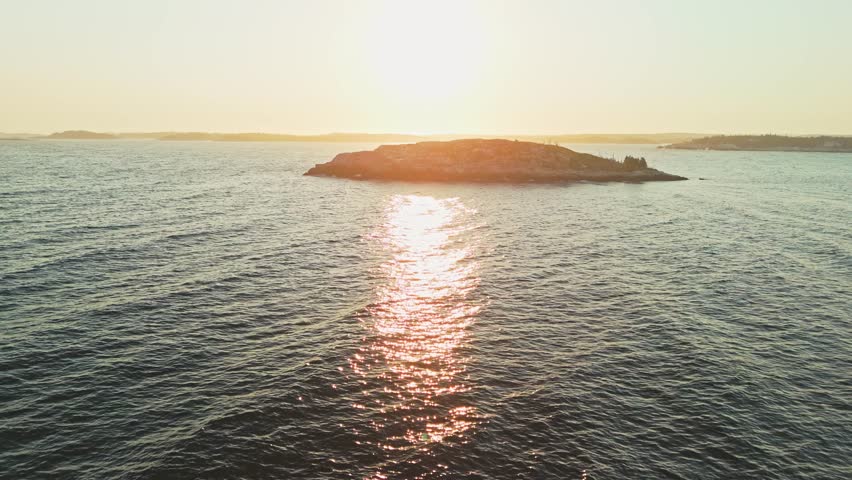 A cinematic drone captures the sunset over the ocean and rocky forest shore in Nova Scotia, Canada. The warm hues highlight the rugged coastline and lush forest. 