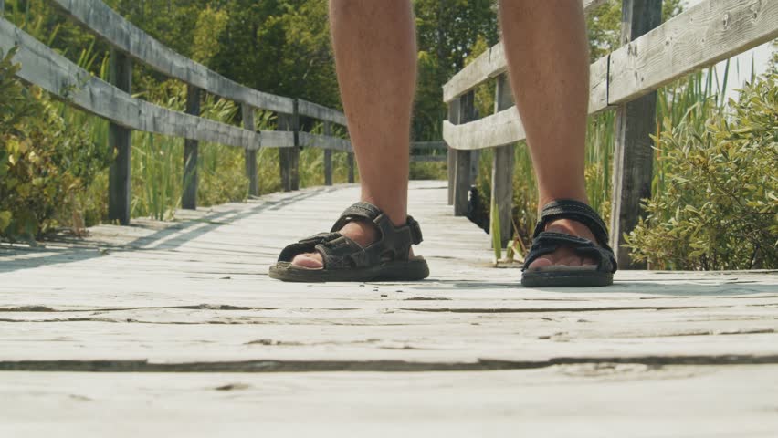 A man stands near a lake in bright sunlight, spraying mosquito repellent on himself. The clear sky and serene waters make the scene idyllic.