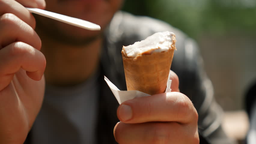 Close up view of ice cream cone, man eating ice cream with spoon on sunny bright hot day.