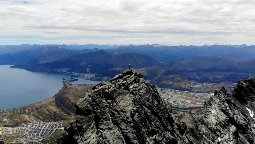 Mountaineer standing ontop of high rocky mountain overlooking Queenstown and Lake Wakatipu in New Zealand.