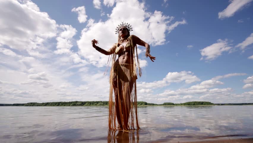 A woman stands in the water, against the background of a lake and a sky with clouds, in an exotic costume with a fringe and a crown, dancing, creating a feeling of freedom and lightness.