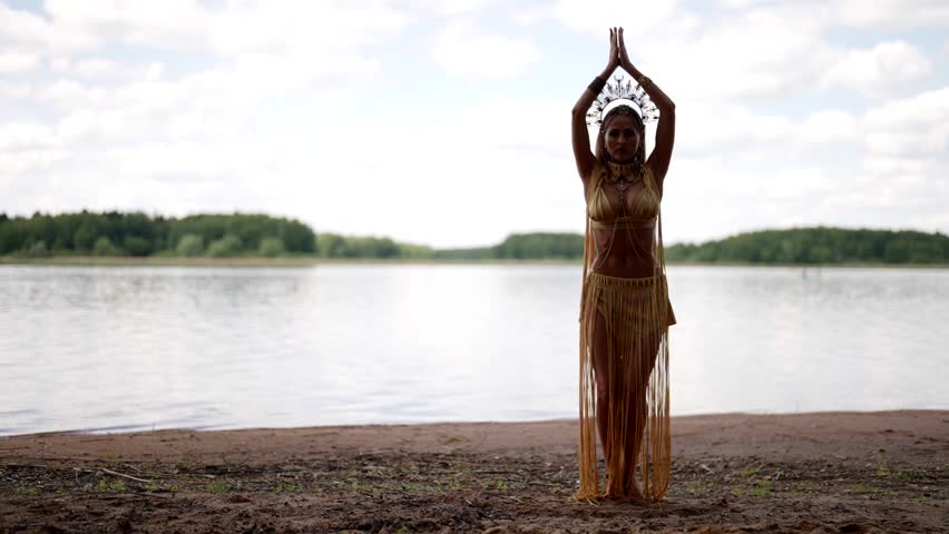 Against the background of a picturesque lake and a blue sky with clouds, a woman in an exotic costume with a crown is dancing, her movements are light and graceful.