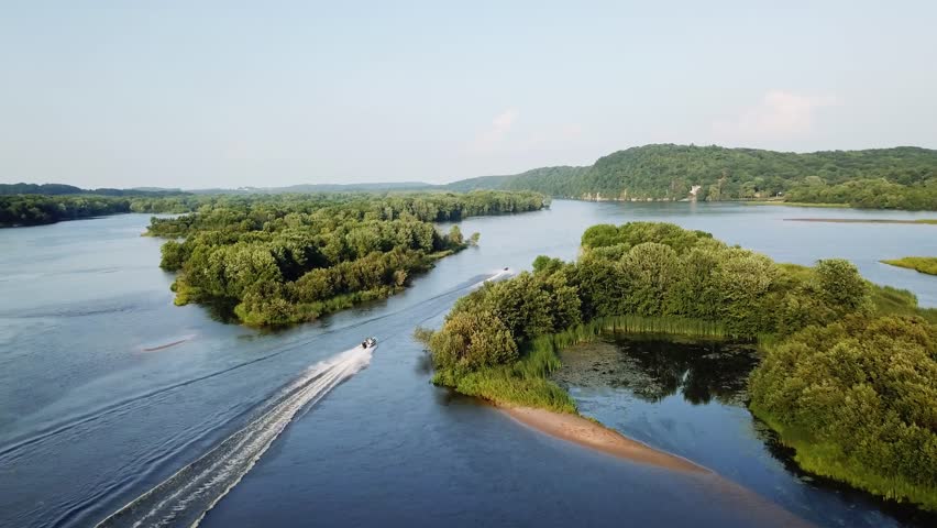 A speedboat cruising down the Wisconsin River, surrounded by lush green forests and rolling hills under a clear blue sky.