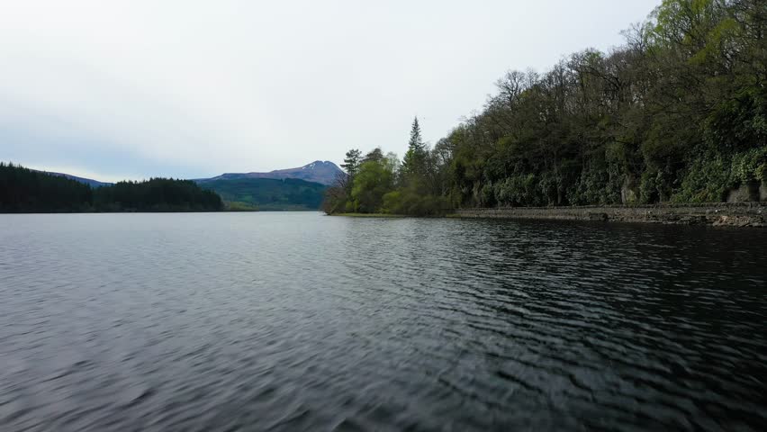 Low aerial flight over water of Loch Ard, green pine forest and road on wooded shoreline, Scottish Highlands, Scotland