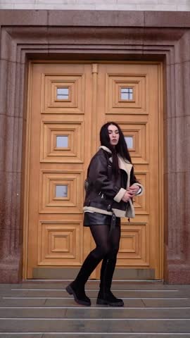 A beautiful brunette girl with dark hair in a jacket looks at the camera while walking in the city