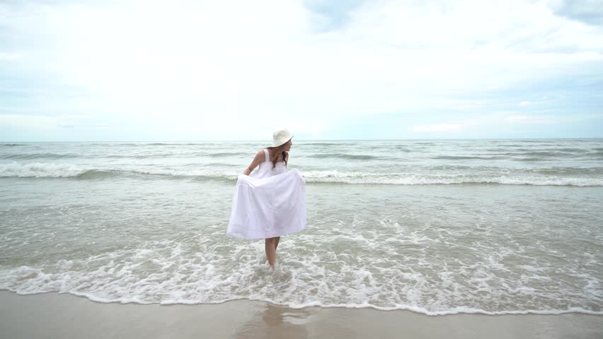 Beautiful Asian girl happy free wearing white dress walking leisurely on the beach and running in the sea waves relaxing summer holiday
