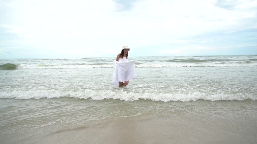 Beautiful Asian girl happy free wearing white dress walking leisurely on the beach and running in the sea waves relaxing summer holiday