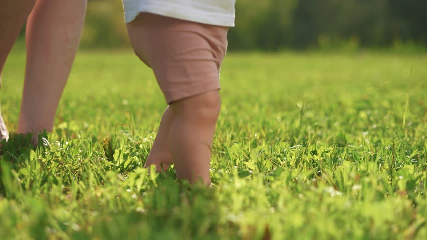 A baby is taking the first steps in the grass. Child walk mother concept. Mom and baby toddler taking first steps. Close-up baby bare feet walking on the grass lifestyle in the park.