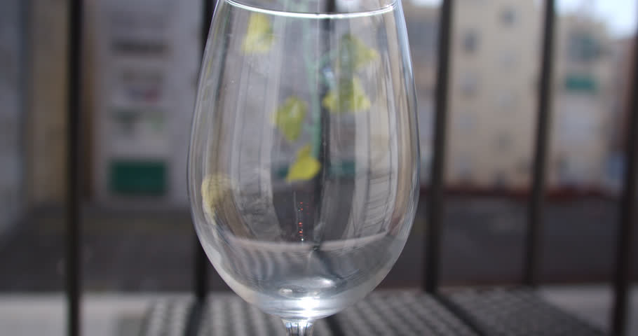 eautiful slow motion shot showing how a couple of ice cubes are placed inside a glass cup on a terrace in the sunlight. There are buildings in the background out of focus.