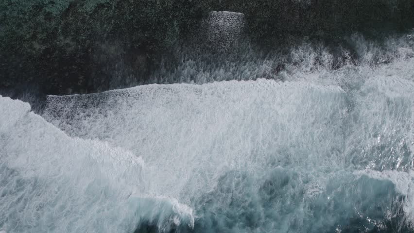 An aerial zooming in view of foamy ocean waves crashing on a cliff on the coast
