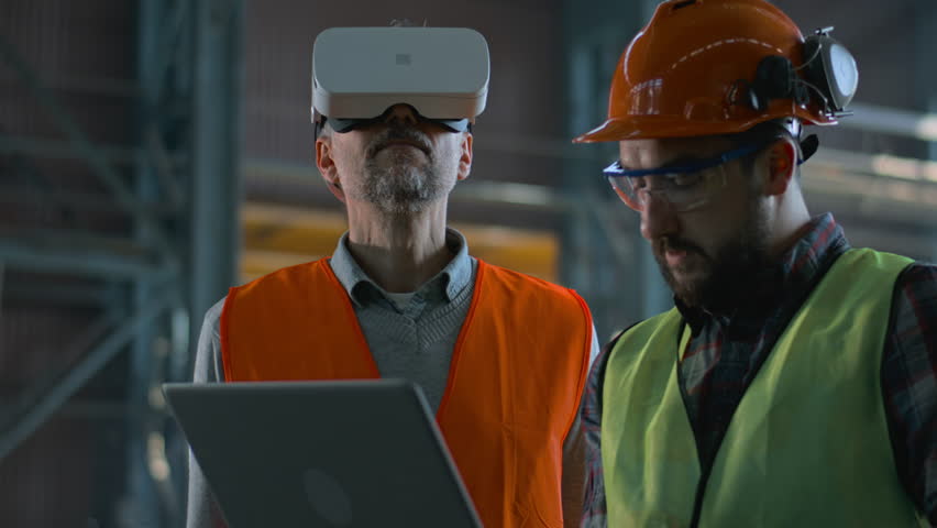Two professional industry technicians wearing safety uniform and hardhats stand at manufacturing factory, use laptop and AR headset. Mature engineer and adult employee work in warehouse. Slow motion. - Powered by Shutterstock - Get 15% off with code: PIKWIZARD15