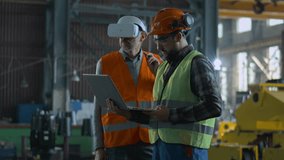 Two professional industry technicians wearing protective uniform and helmets stand at manufacturing factory. Mature engineer uses VR headset, adult employee work in laptop computer. Slow motion. - Powered by Shutterstock - Get 15% off with code: PIKWIZARD15