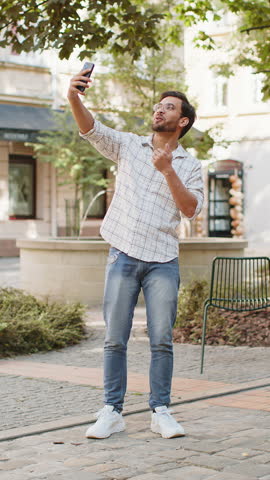 Cheerful Hispanic man blogger taking selfie on smartphone, communicating video call online with family friends, recording stories for social media vlog, waving hello. Arabian guy on urban city street.