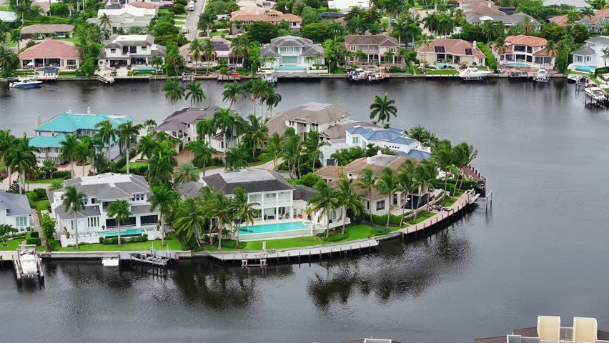Waterfront homes with private docks in Florida. Lush greenery and palm trees surround luxury properties. Canal view, aerial.