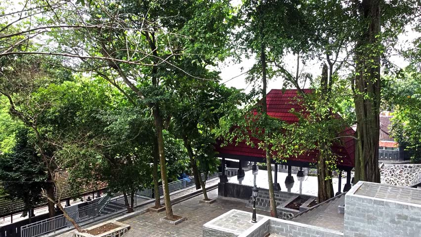 A serene park scene featuring lush green trees, a traditional red-roofed pavilion, and stone pathways. The area is well-maintained, inviting visitors to relax and enjoy nature.