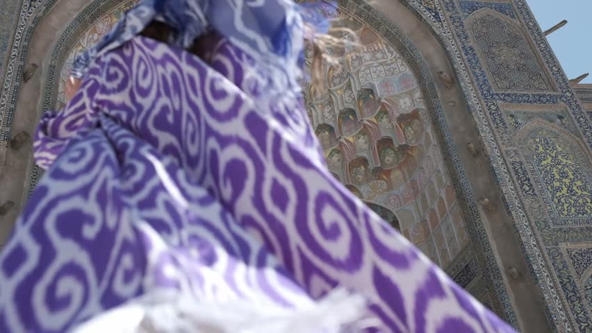 A young woman with long hair in an oriental robe and skullcap stands in the wind near an ancient mosque