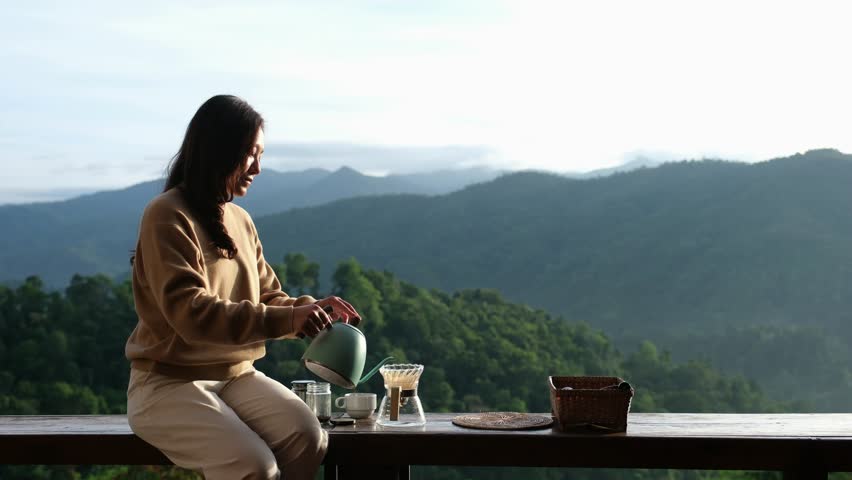 A woman sitting on wooden balcony and making drip coffee with a beautiful mountain view