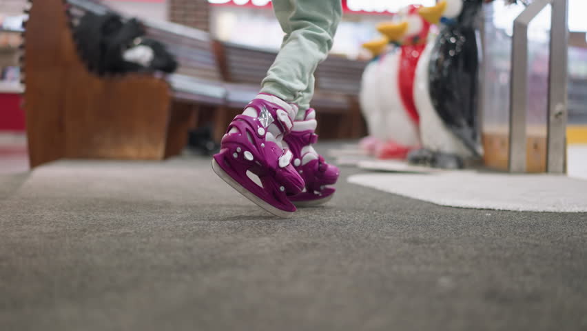 Close-up of a person walking into an ice rink wearing purple skates and ash-colored trousers, with blurred view of people skating in the background