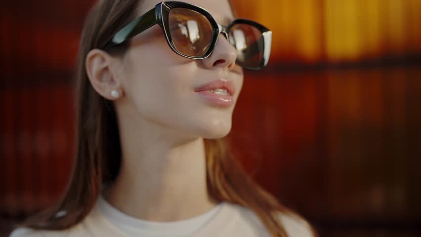 A fashionable young woman with long brown hair wears large sunglasses, basking in warm sunlight with a thoughtful look, against a vibrant urban backdrop. Stylish Young Woman in Sunglasses Enjoying