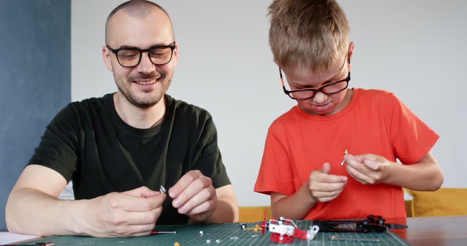 Happy father teaches son to screw metal details of constructor set at table. Man and toddler boy with glasses assemble model together at home