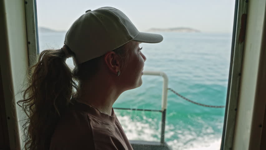 Woman in casual wear enjoys sea view from ferry door. Female gazes at ocean horizon, standing on boat. Calm maritime journey, tranquil travel scene. Relaxed tourist watches water, islands in distance.