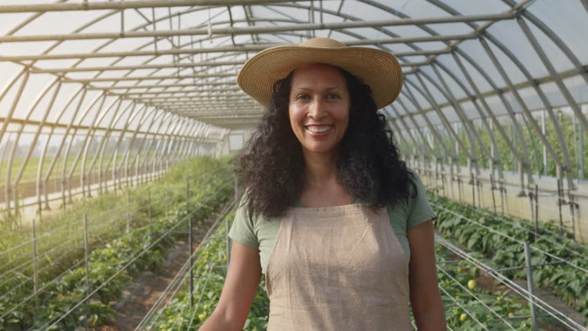Smiling latin woman standing in greenhouse portrait, front view medium shot. Cheerful female hothouse employee wearing apron and straw hat looking at camera