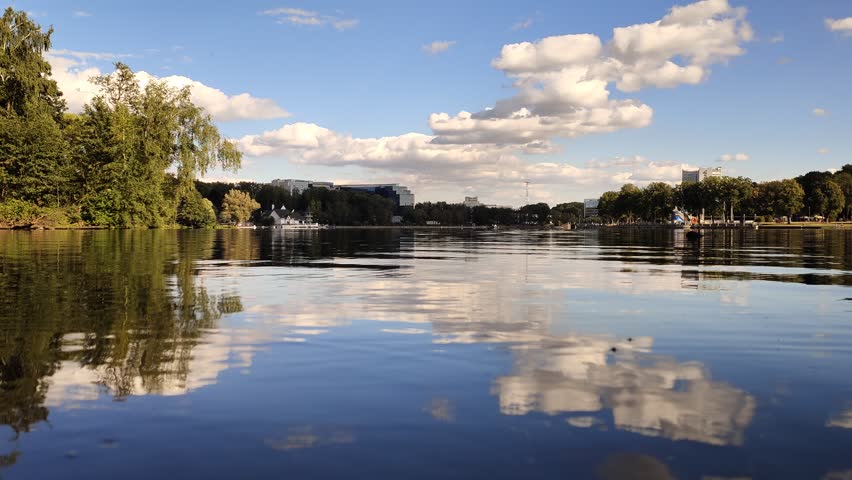 Birch trees and other trees grow on the shores of the lake. On the far shore - the embankment, TV tower and city buildings. The water is rippling. Blue sky with clouds reflected in the water. Sunny