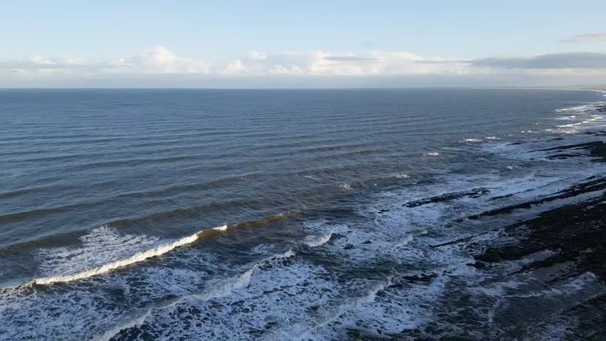 A descending drone over green highlands by splashing sea waves in Caithness, Scotland