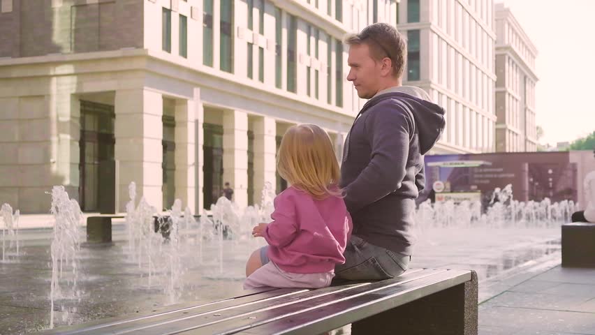 Cute Caucasian child girl toddler with her father sitting near the fountain.