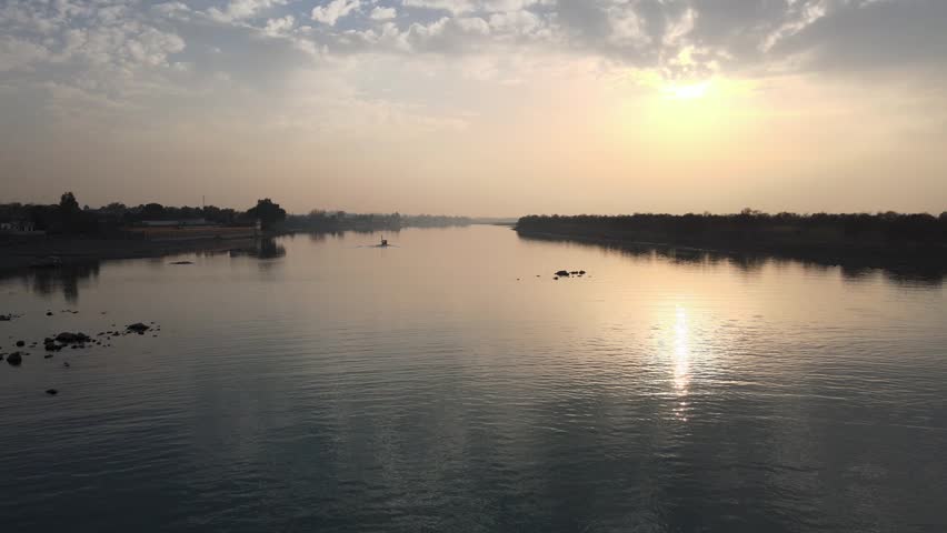 An aerial shot over a river between rural fields under shiny sunset sky reflecting on the water