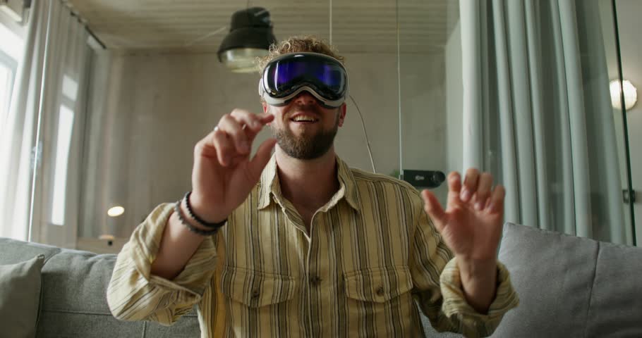 A man in a virtual reality headset is typing on a virtual keyboard and moving his hands sitting on the couch. Daylight, home interior