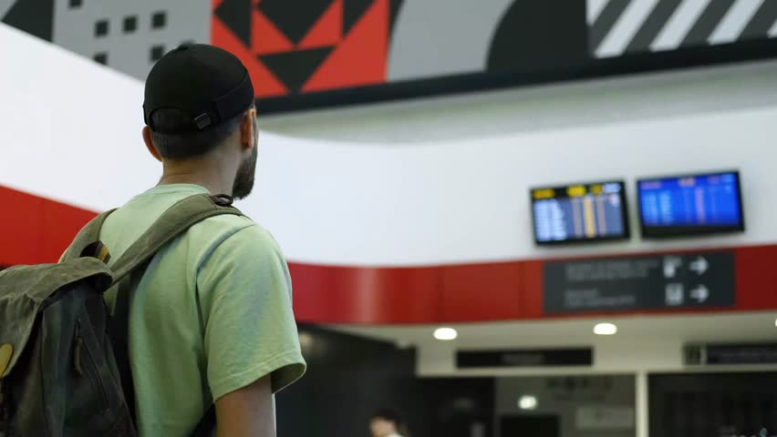 Young man at the airport looking at the list of destinations, rear view. Tourist guy with green retro backpack waits for a train at a train station. A new railway station in Belgrade, Serbia