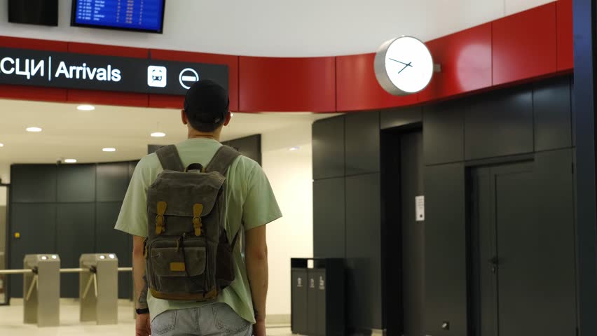 Young man at the airport looking at the list of destinations, rear view. Tourist guy with green backpack waits for a train at a train station. Belgrade, Serbia