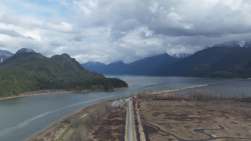 A drone view above a road between dry fields surrounded by water and mountains on a cloudy day